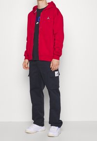 Felpa rossa con zip e tasche frontali, maglietta nera, pantaloni cargo blu con tasche laterali e sneakers bianche con accenti blu.