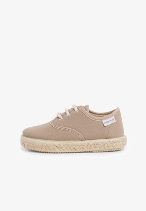 Chaussure en toile beige à lacets avec semelle en jute tissée et une petite étiquette en tissu cousue près du talon sur un fond blanc.