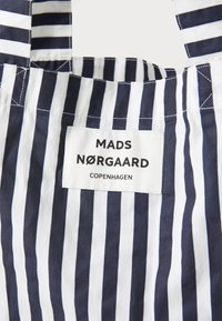Csíkos tote táska váltakozó sötétkék és fehér mintákkal. Fényes felirattal ellátott textilcímke, melyen a "MADS NØRGAARD COPENHAGEN" felirat olvasható fekete színben.