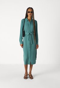 Abito camicia turchese con maniche lunghe, chiusura a bottoni e cintura da annodare. Caratterizzato da una lunghezza midi e fessure laterali, abbinato a sandali neri.