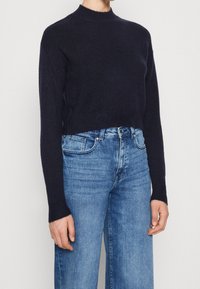 Marineblå kortärmad tröja med hög hals och långa ärmar, tillverkad av strukturerat stickat material, kombinerad med högmidjade ljusblå jeans.