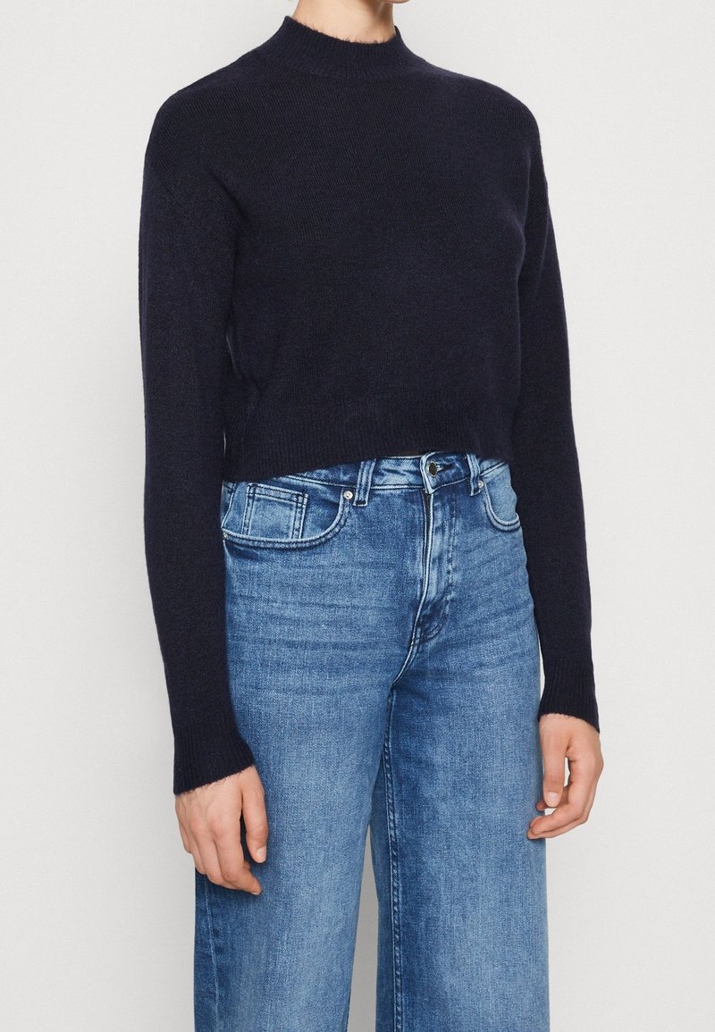 Marineblå kortärmad tröja med hög hals och långa ärmar, tillverkad av strukturerat stickat material, kombinerad med högmidjade ljusblå jeans.