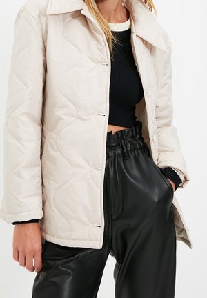 Kobieta w beżowej pikowanej kurtce, na czarnym krótkim topie i czarnych, skórzanych, wysokich spodniach z paskiem typu paperbag, z ręką w kieszeni.