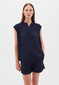 Mörkblå ärmlös blus med knäppning, veckad halsringning och mjukt draperad tyg, ihopparad med matchande mörkblå shorts.