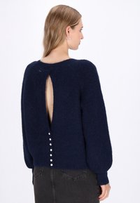 Marineblauer Strickpullover mit fuzzie Textur, langen Ärmeln und einem Cut-out am Rücken. Verfügt über eine Reihe weißer Knöpfe entlang des unteren Rückens.