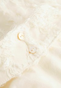 Crèmewitte stof met rafelige randen en twee ronde, lichtgekleurde knopen bij de kraag. Zachte textuur en gladde afwerking.