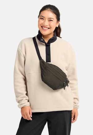 Lächelnde Frau in beigefarbenem Fleecepullover mit schwarzem Kragen und schwarzer Umhängetasche steht vor einfarbig hellem Hintergrund.