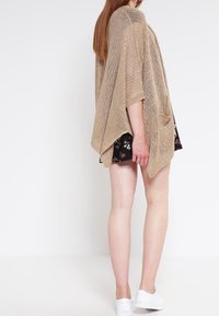 Lätt beige stickad poncho med öppen design, korta ärmar och avslappnad passform. Kombineras med en blommig svart klänning och vita sneakers.