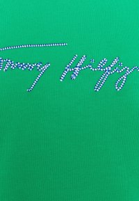 Chemise en coton verte avec un logo brodé en texte rayé bleu. Le tissu a une texture lisse et un design ajusté.