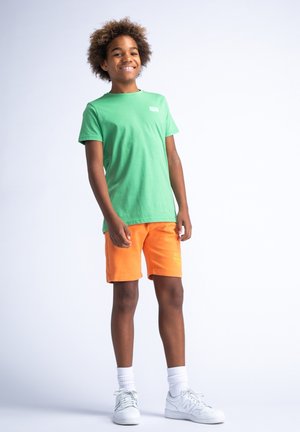 Camiseta de algodón verde con mangas cortas, combinada con pantalones cortos naranjas que tienen un logotipo de la marca. Zapatillas blancas con acentos grises.