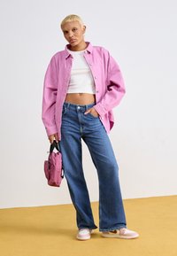 Różowa koszula z sztruksu, biały krótki top, niebieskie szerokie spodnie dżinsowe i różowe trampki. Trzyma małą różową torebkę. Jednolite tło z żółtą podłogą.