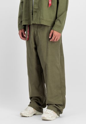 Unterer Teil einer Person, die weite olivgrüne Hosen, eine passende Jacke mit roten Etiketten und weiße Mesh-Sneaker vor grauem Hintergrund trägt.