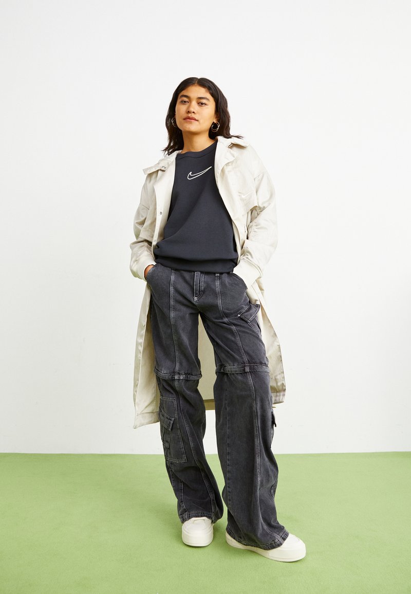 Trench coat de gran tamaño en color crema, sudadera azul marina con el logo blanco de Nike, pantalones cargo oscuros con bolsillos, zapatillas blancas, de pie sobre un suelo verde.