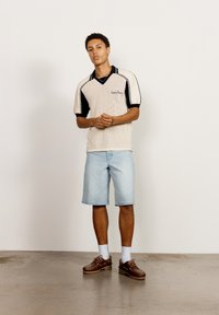 Kortermet strikket polo i krem med svarte detaljer, kombinert med lyseblå denimshorts. Modellen har på seg brune sko og hvite sokker.