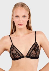 Bralette triangular de encaje negro con paneles de malla transparente, tirantes finos y un acento decorativo de hardware dorado en el centro frontal.