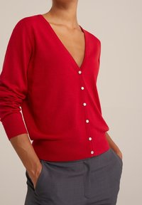 Röd V-ringad kofta med lång ärm och detaljer i silverknappar. Tillverkad av mjukt stickat material, med en avslappnad passform och ribbade ärmslut.