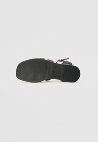 Sort sandal med flad sål, der har en glat læderoverdel, justerbar ankelrem og minimale hardwaredetaljer.