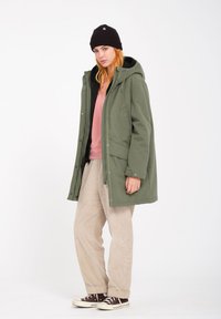 Parka verte avec une capuche, fermetures à glissière et à pression, coupe surdimensionnée, superposée sur un pull rose. Pantalons en velours côtelé beige et baskets noires.