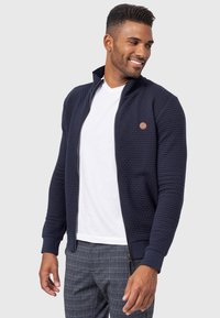 Mørkeblå zip-up jakke i tekstureret stof, rib-strikkede ærmer og kant samt en lille logo detalje. Båret over en hvid t-shirt og mønstrede bukser.