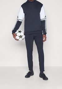 Navy Trainingsanzug mit weißen Akzenten, bestehend aus einem Kapuzenpullover und schmal geschnittenen Hosen. Das Model hält einen Fußball mit schwarzen und grünen Details.