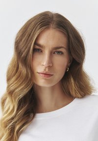 Dlhé vlnité vlasy, neutrálny odtieň pleti a jemný make-up. Na sebe má bielu košeľu a jednoduché zlaté kruhové náušnice. Neutrálny pozadia.