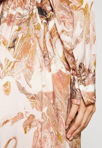 Blusa in tessuto astratto marmorizzato rosa e beige, con maniche lunghe, polsini abbottonati e texture liscia. Accenti dorati notevoli.