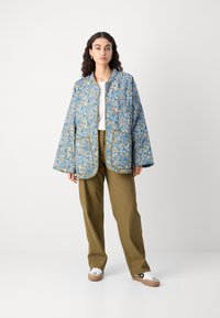 Giacca trapuntata blu con motivo floreale, maniche lunghe e rifiniture dorate, dotata di due tasche frontali, abbinata a pantaloni khaki e sneakers bianche.