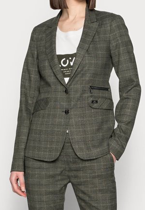 Kobieta ubrana w dopasowaną, szarą kratkowaną marynarkę z zapięciem na guziki oraz dopasowane spodnie, do tego biała koszulka z grafiką pod spodem.