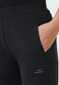 Schwarze Sporthosen mit einem enganliegenden Bund, seitlichen Taschen und einem kleinen "VENICE BEACH"-Logo am unteren linken Bein. Glatte Stoffoberfläche.
