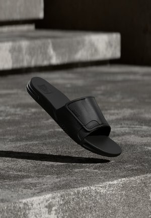 Sandalo nero a ciabatta con plantare testurizzato e larga fascia regolabile, posizionato in aria sopra gradini di pietra in luce soffusa.