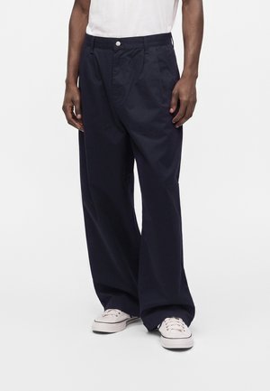 Persona que lleva pantalones anchos azul marino oscuro, zapatillas blancas y una camisa blanca, de pie contra un fondo blanco liso.