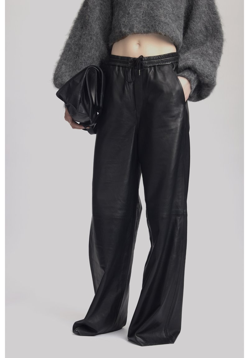 Personne portant un pull court gris, un pantalon en cuir noir à jambes larges, tenant un sac en cuir noir plié dans une main.
