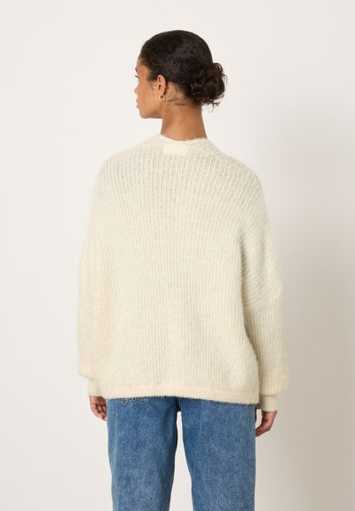 Pull en crochet crème à coupe ample, épaules tombantes, texture côtelée et ourlet roulé, associé à un jean bleu taille haute.
