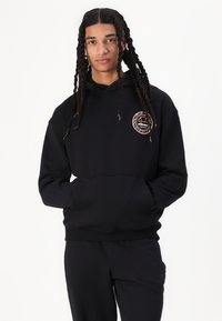 Svart hettegenser med et rundt logo som har et oransje basketballdesign. Stoffet er mykt med en jevn tekstur, og den har en frontlomme i kengurustil.