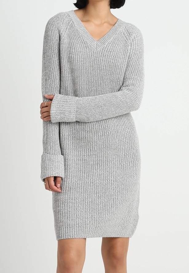 Femme portant une robe-pull en maille côtelée gris clair avec des manches longues à poignets et un col en V, debout devant un fond blanc uni.