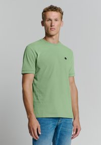 Groene katoenen T-shirt met korte mouwen en een ronde hals, voorzien van een klein zwart logo op de linkerkant van de borst. Gemaakt in combinatie met blauwe jeans.
