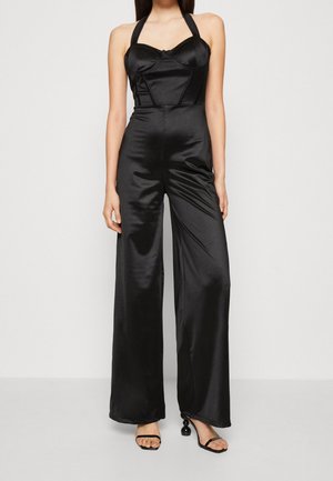 Kvinde iført en sort satin halter-neck jumpsuit med vide ben og sorte åbne tå-hæle med dekorative runde hæle.