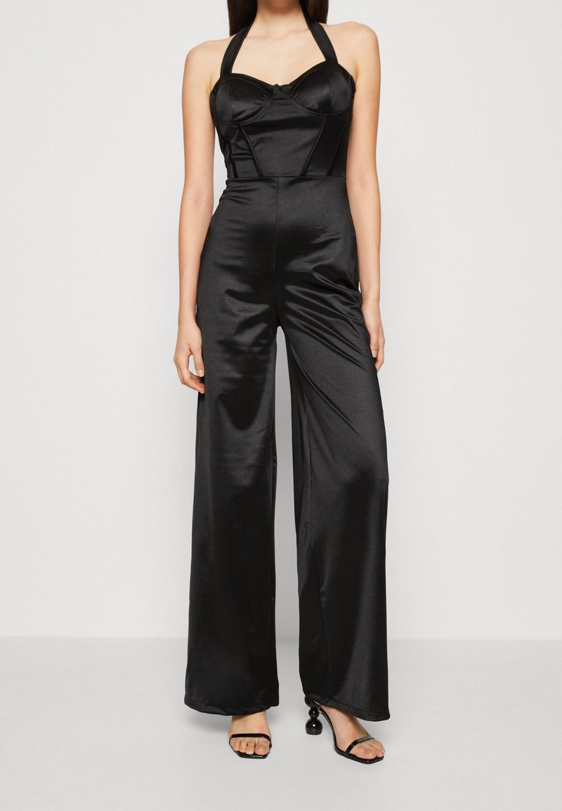 Femme portant une combinaison noire en satin à col halter avec jambes larges et talons ouverts noirs avec talons ronds décoratifs.