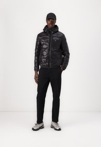 Giubbotto imbottito nero con cappuccio, texture lucida, zip sul davanti, tasca sul petto, abbinato a pantaloni neri e scarpe grigie con suola texturizzata.