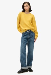 Geel gebreide trui met een losse pasvorm, gedragen met vervaagde blauwe wijde broek en zwarte veterschoenen, die een casual en ontspannen uitstraling laat zien.