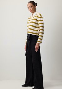 Gestreepte cardigan in crème en olijfgroen breiwerk, gecombineerd met hoge zwarte broeken met een strikceintuur, gedragen met puntige zwarte schoenen.