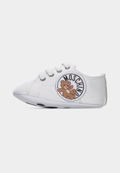 Λευκό παιδικό παπούτσι με δέσιμο κορδονιών και στρογγυλό σήμα που απεικονίζει ένα καφέ αρκουδάκι και το κείμενο "MOSCHINO" στο πλάι.