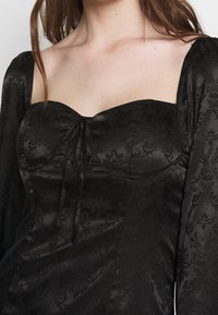 Černý strukturovaný top se čtvercovým výstřihem, dlouhými nabíranými rukávy a detailem vázání. Tkanina má jemný květinový vzor a saténový vzhled.