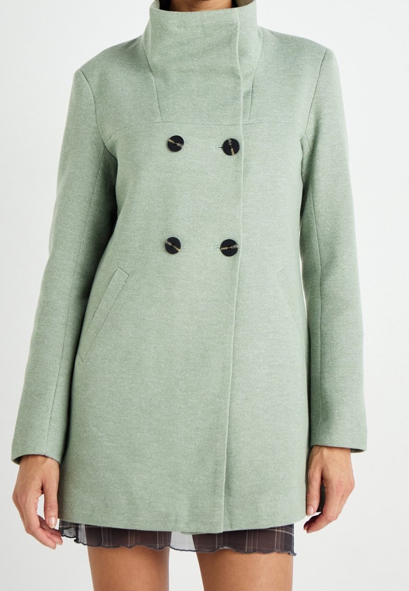 Abrigo verde de corte doble, con cuello alto, fabricado en tejido texturizado, con botones negros y bolsillos laterales.