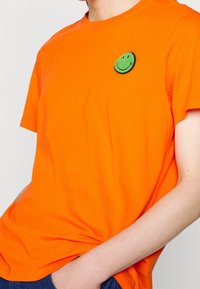 Personne portant une chemise à manches courtes orange vif avec un petit patch de smiley vert sur la poitrine gauche en haut.