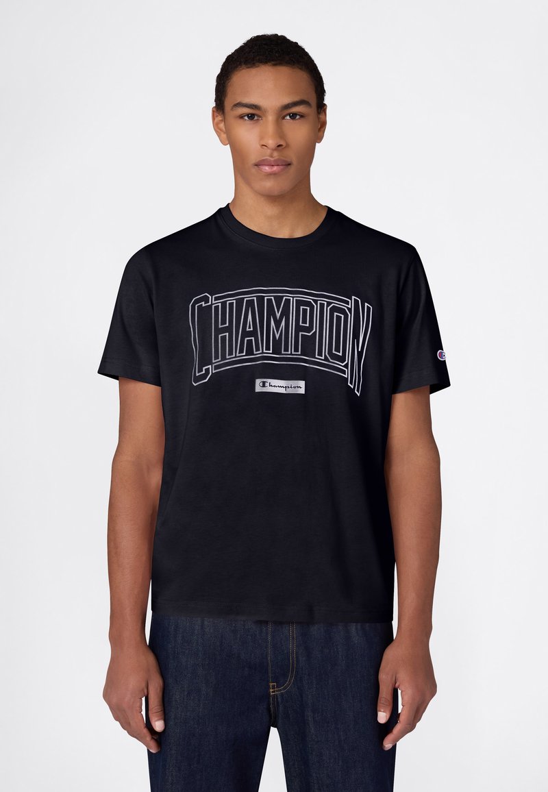 T-shirt en coton noir à manches courtes, avec un logo blanc "CHAMPION" sur la poitrine et une petite étiquette près de l'ourlet.