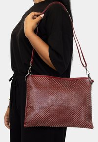 Bolso de hombro de cuero burdeos con acentos de tachuelas, cierre con cremallera y correa ajustable. Textura suave, forma rectangular y acabado brillante.