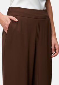 Brązowe spodnie typu wide-leg o gładkiej teksturze i wysokim pasie. Posiadają boczne kieszenie oraz plisy dla dodatkowego efektu.