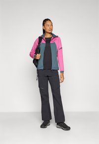 Fleece jacket in pink and teal with zip front, paired with black pants and hiking boots; model carries a black backpack.