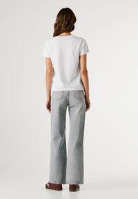 Žena s dlouhými hnědými vlasy stojí zády, obléká bílé tričko, světle šedé široké džíny a bordó tenisky.